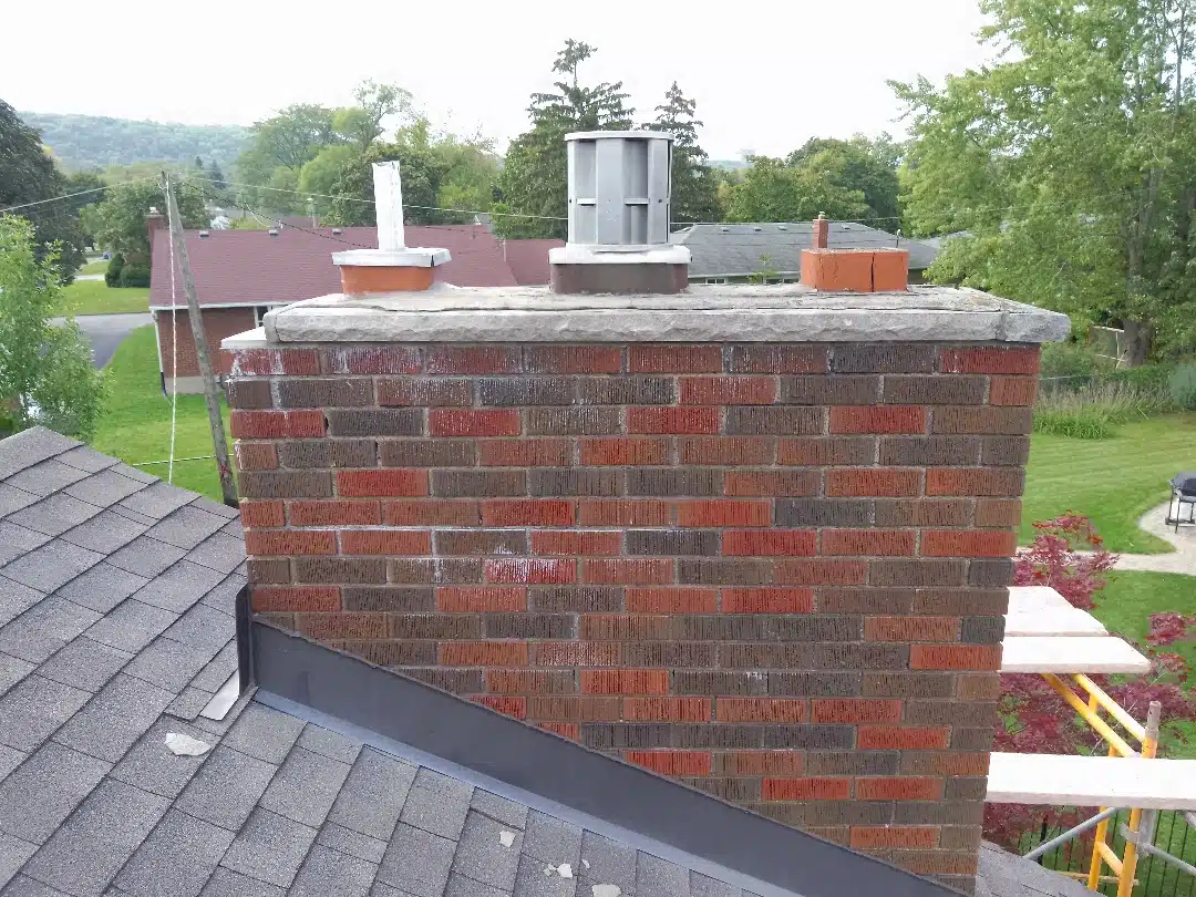 This image shows a brick chimney or chimney-like structure on the roof of a building The chimney is made of red and gray bricks with a metal or concrete cap on top The structure is surrounded by a sloped roof likely made of asphalt shingles and there are trees and greenery visible in the background suggesting a residential or rural setting